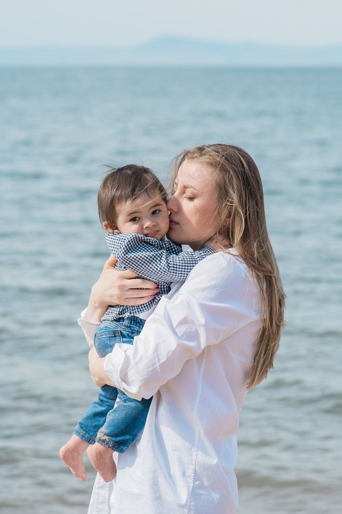 A loving mother holding and kissing her baby boy on a sunny beach day, embracing family joy.