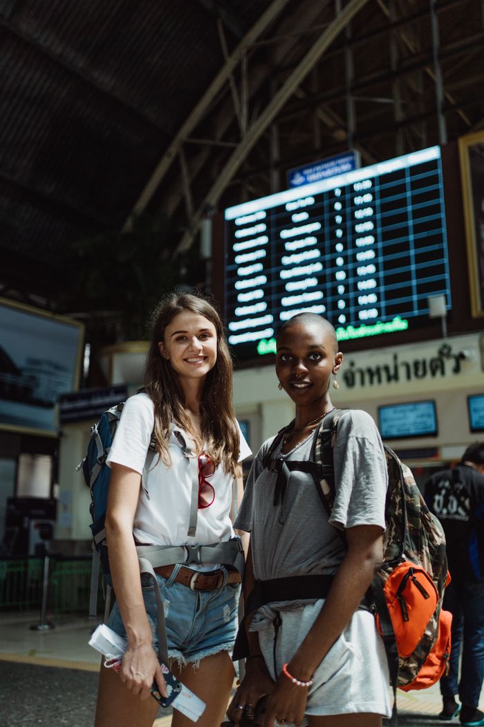 Two women with backpacks smiling in a train station, ready to travel.
