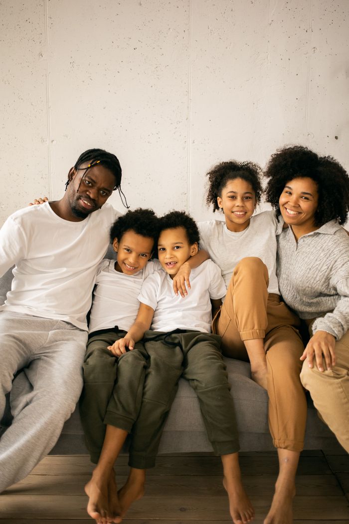 Delighted barefoot African American family with kids wearing casual wear smiling and looking at camera while sitting on comfortable couch