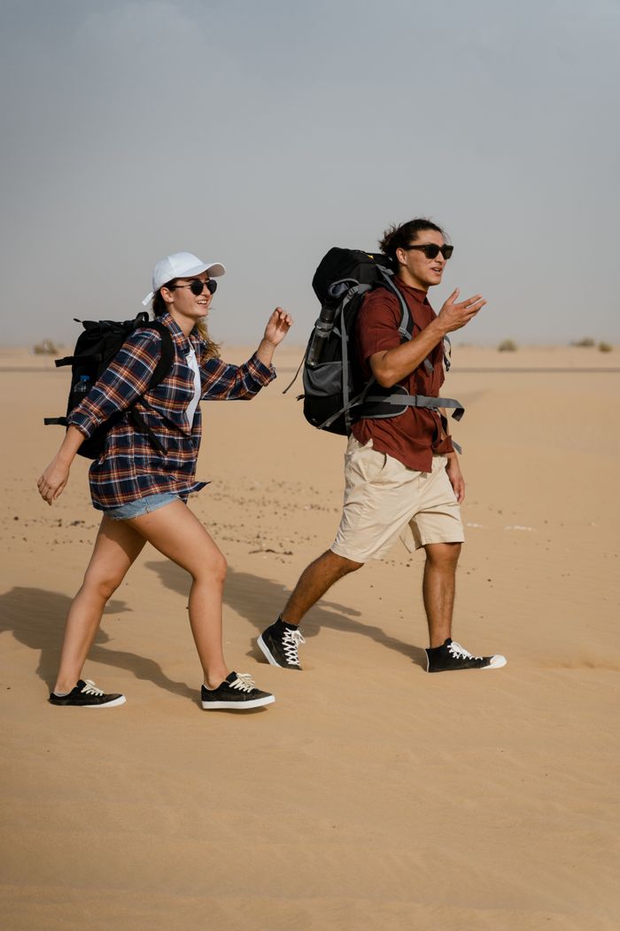 Two young adults hiking with backpacks across an open desert on a sunny day, embracing adventure.